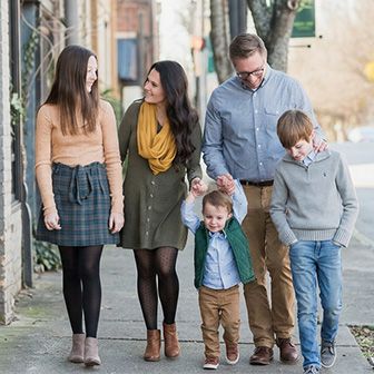 A family of five walks side by side on a sidewalk, with buildings on one side and trees and a street on the other.