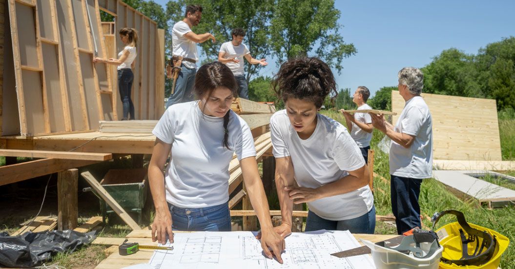 A picture of two adults standing over house blueprints held up by a board and saw horses while others in the background build the frame of a house.