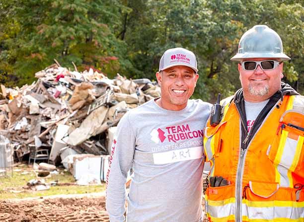 Two smiling Team Rubicon disaster response workers at a debris site, one in gray shirt and cap, one in orange safety vest.