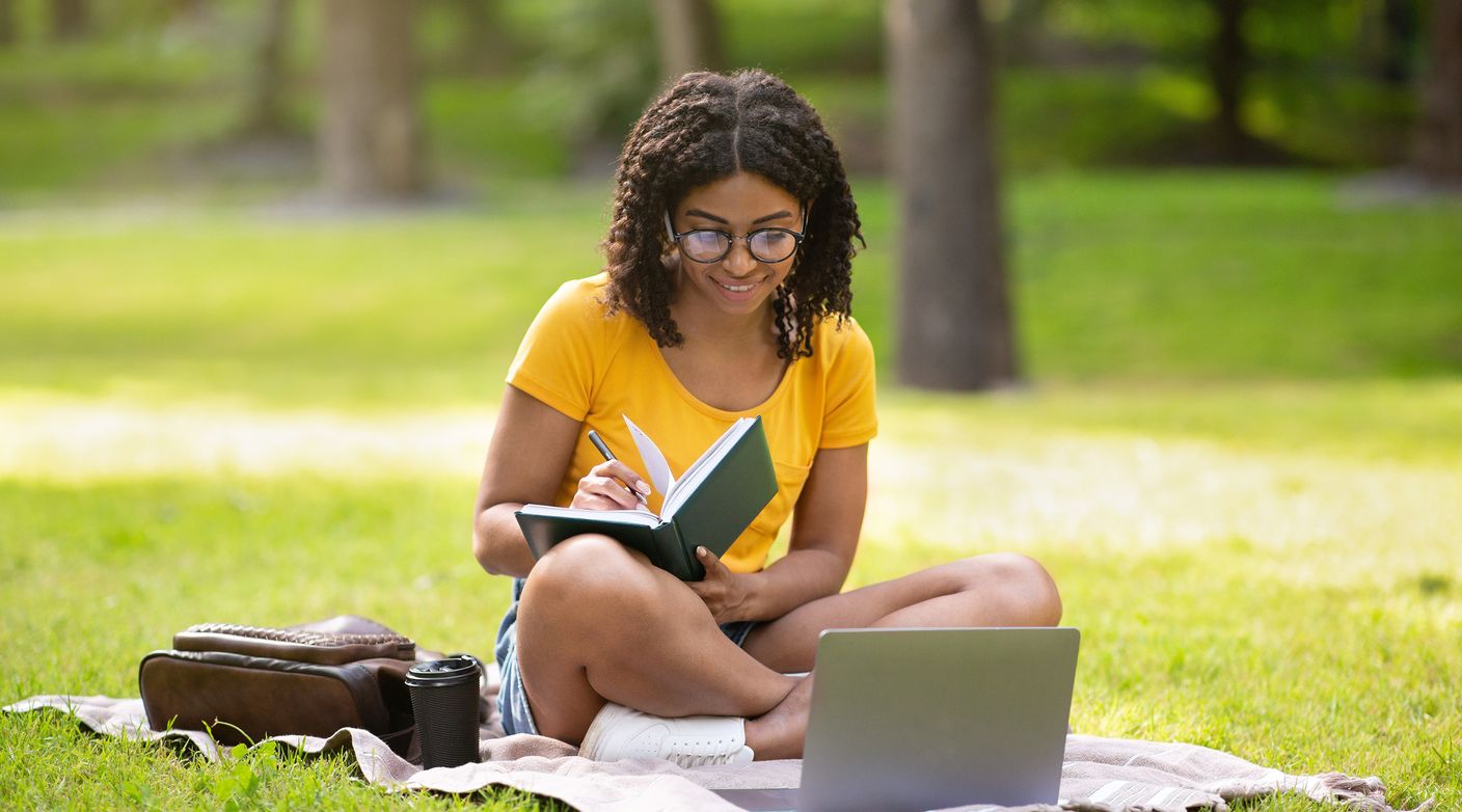 Young adult female is taking notes while sitting in a park watching her laptop.