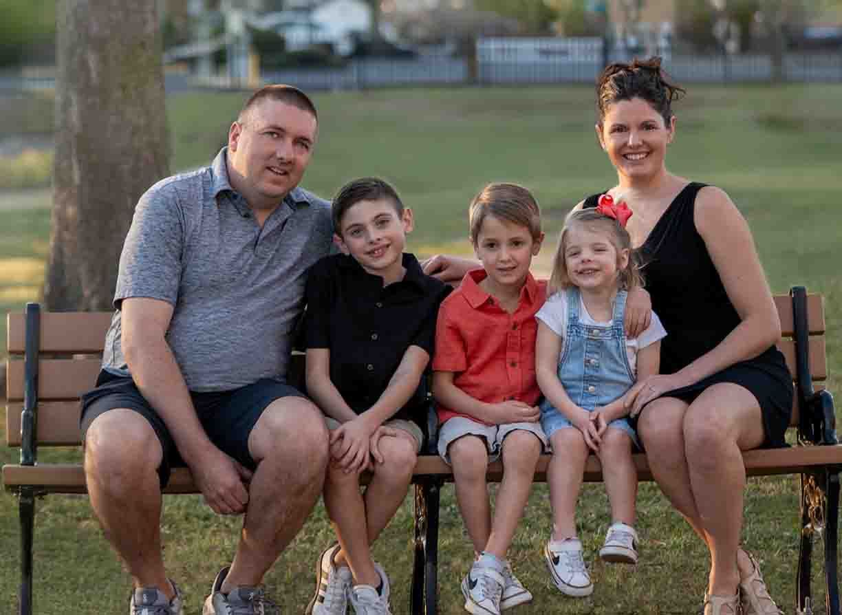 A family of five sitting on a park bench, including two parents and three children, smiling at the camera outdoors.