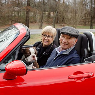 Retired couple driving in red convertible with their dog.