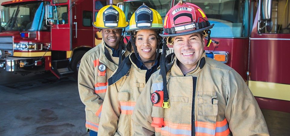 Three smiling firefighters standing in front of fire trucks.